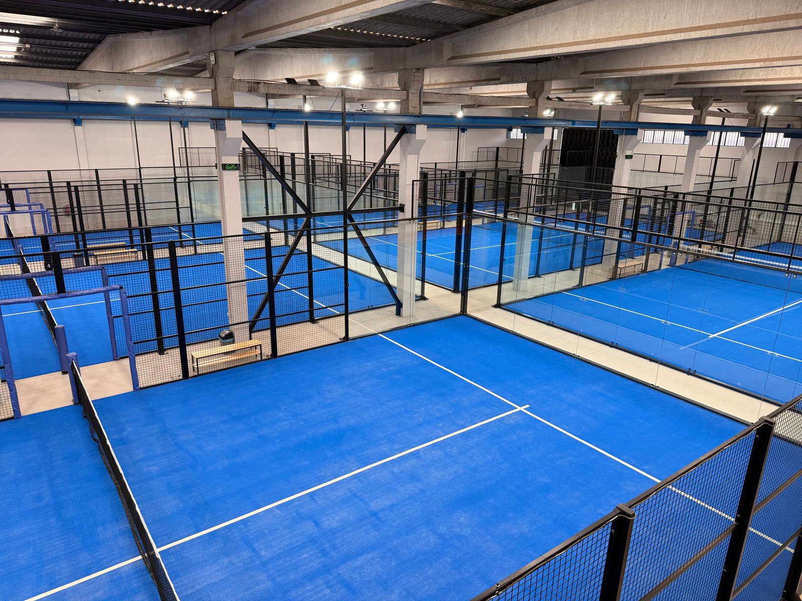 Quadras de badminton internas com piso azul, cercadas por telas de proteção, em uma instalação esportiva com teto de concreto e iluminação.