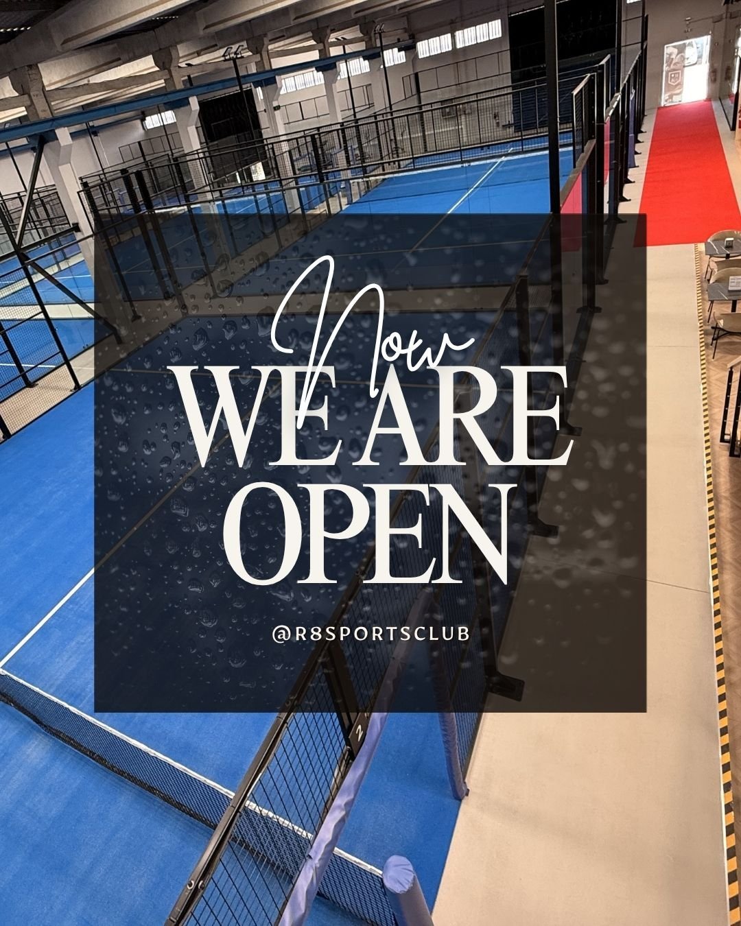 Pista de tênis de mesa em uma quadra coberta, com barreiras dividindo as áreas de jogo, e uma mensagem que diz "Now We Are Open" com o nome @R8SportsClub ao fundo.
