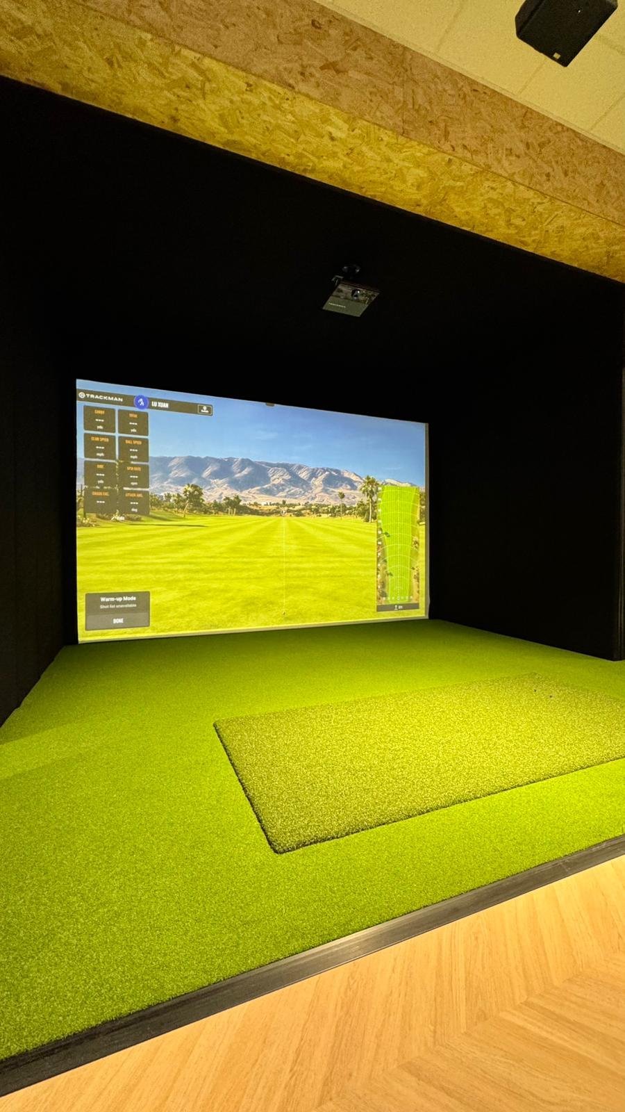 Sala de simulação de golfe com tela grande exibindo um campo de golfe e um simulador de movimento.