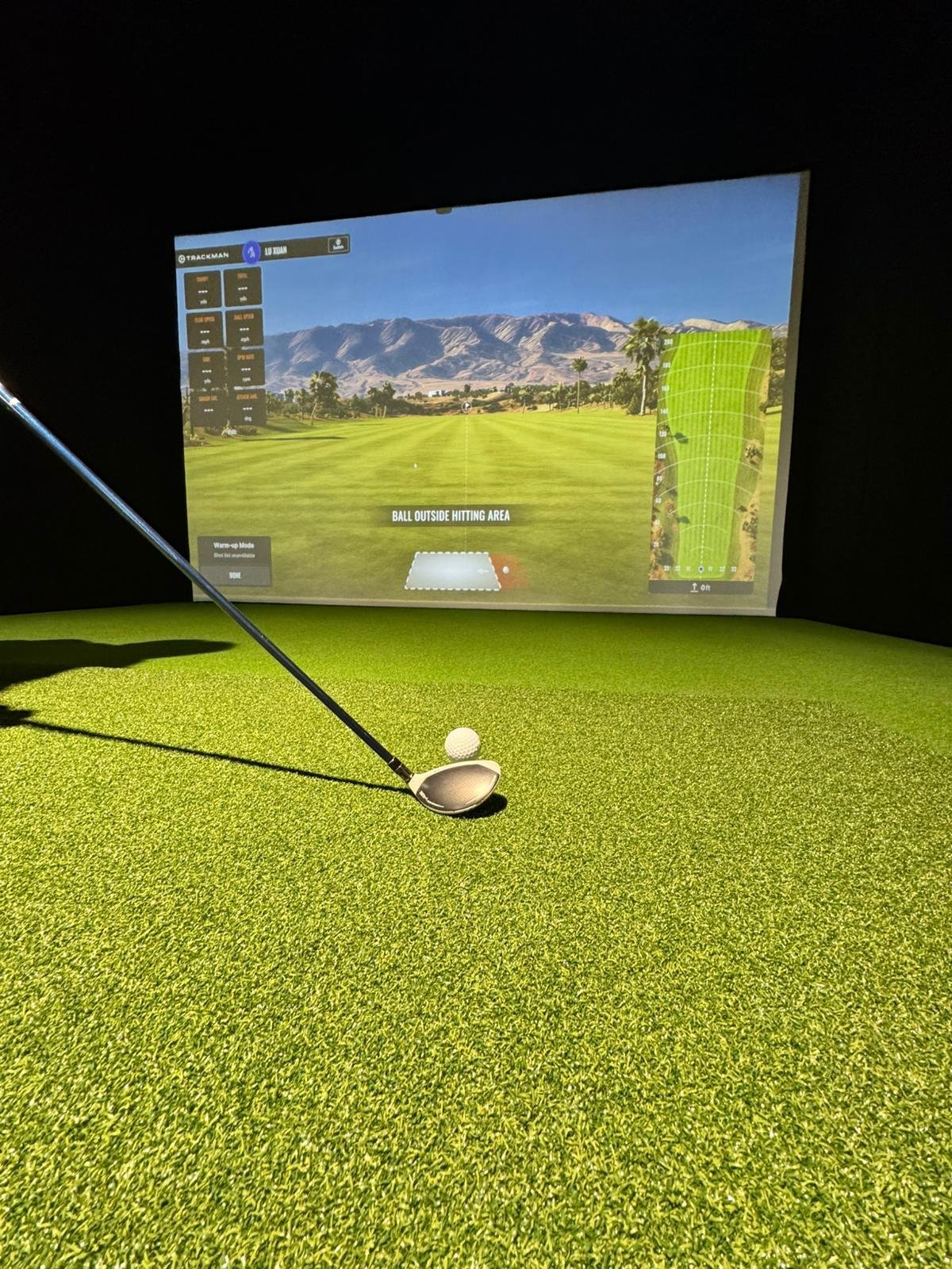 Imagem de uma pista de golfe com uma bola e um taco em uma sala de simulação de golfe, com tela de projeção exibindo um campo de golfe e montanhas ao fundo.