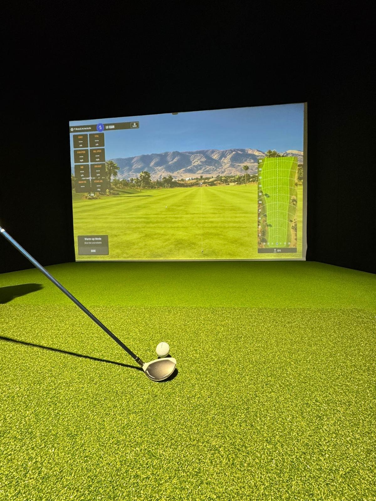 Uma sala de simulação de golfe com uma tela grande mostrando um campo de golfe e um drive de golfe com bola e taco na grama verde artificial.