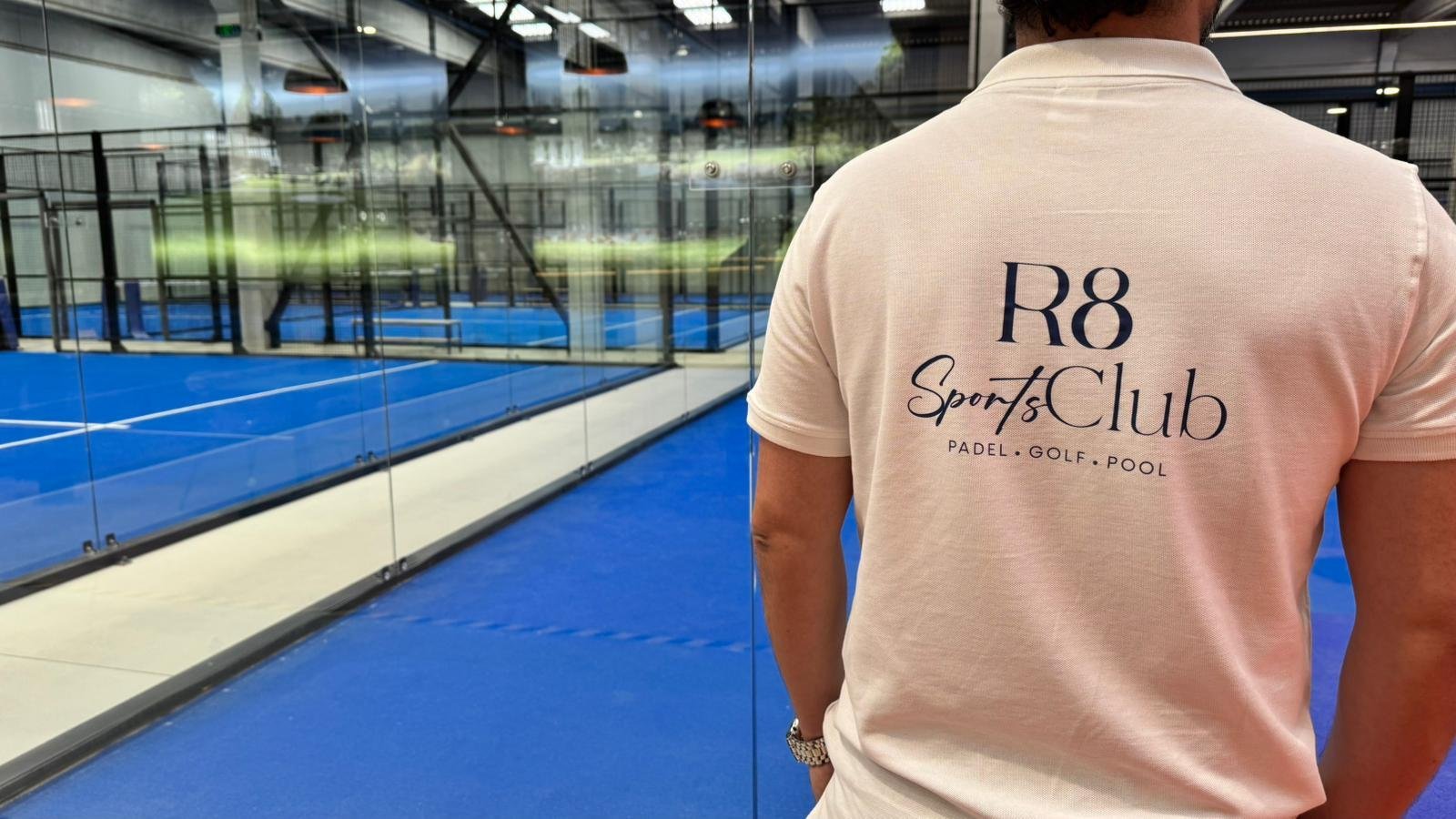 Pessoa com camiseta clara com a inscrição "R8 Sports Club" vista de costas, ao lado de uma quadra de padel coberta com parede de vidro.