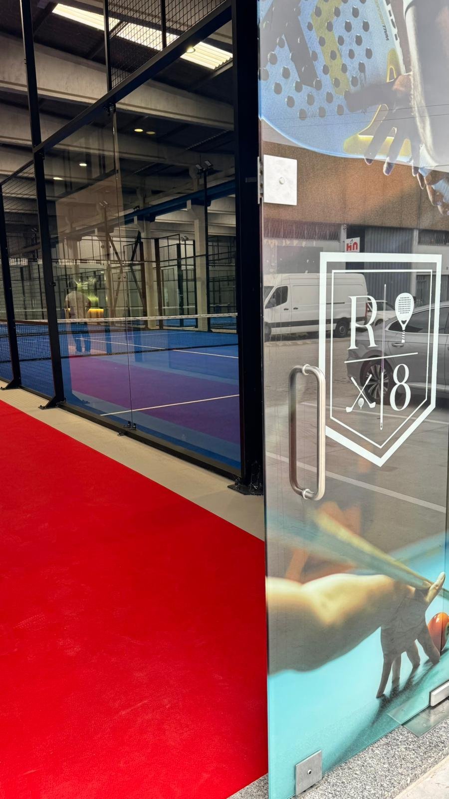 Entrada de uma quadra de Tênis com vidro transparente que possui uma logomarca na porta, exibindo uma raquete, uma bola e o texto 'R X 8'.
