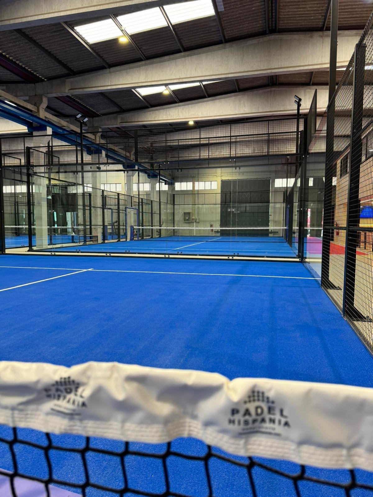 Quadra de padel coberta com piso azul, cercada por grades de metal pretas, com iluminação artificial e janelas ao fundo
