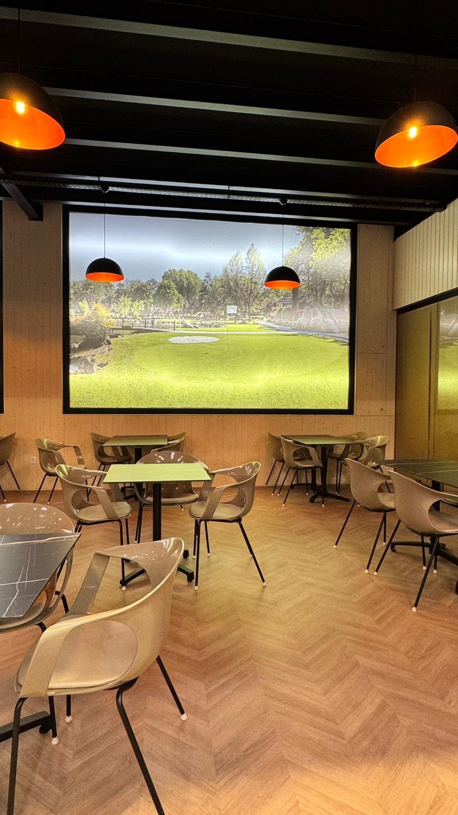 Sala de café ou restaurante com mesas e cadeiras, parede de madeira, luminárias pretas com interior laranja e uma tela grande exibindo um campo de golfe com árvores ao fundo.