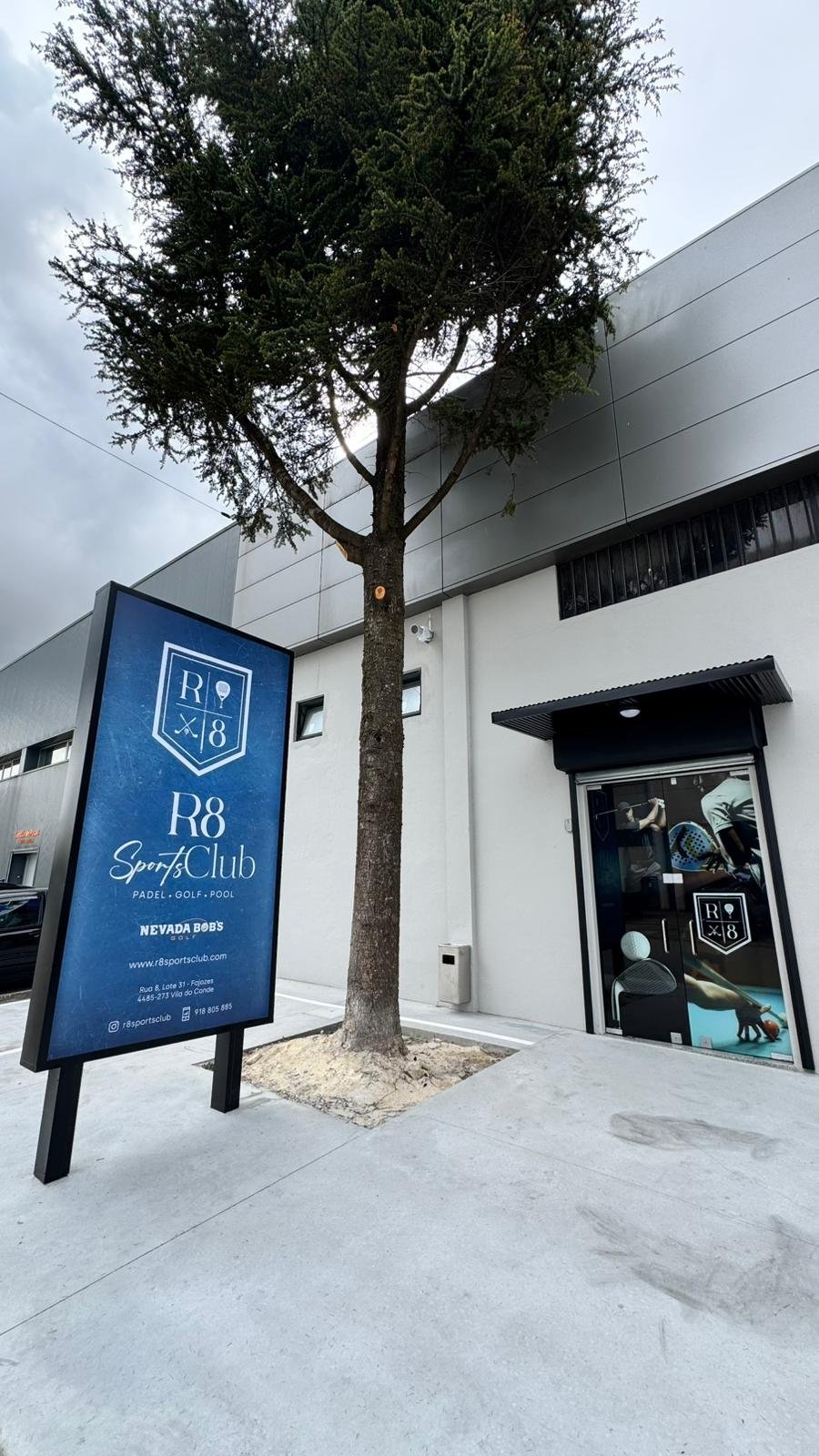 Rua com fachada de prédio branco, uma árvore no meio, uma placa de sinalização azul para clube esportivo de paddle, golfe e piscina, e uma porta de entrada com vidro mostranto pessoas praticando esportes internos.